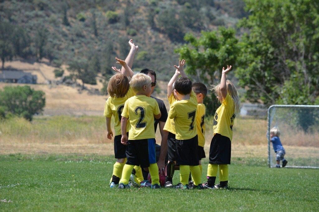 A group of young soccer players in yellow jerseys gathered in a huddle with their coach on a sunny field.
